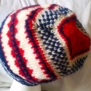 May include: A knitted hat with a red, white, and blue pattern. The hat features a repeating zig-zag design in red and white, with blue accents. The hat is made of a soft, textured yarn.
