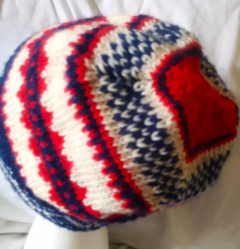May include: A knitted hat with a red, white, and blue pattern. The hat features a repeating zig-zag design in red and white, with blue accents. The hat is made of a soft, textured yarn.