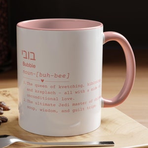 May include: A white ceramic mug with a pink rim and handle. The mug features a definition of the word "Bubbie" in Hebrew and English. The definition reads: "Bubbie noun-[buh-bee] 1. The queen of kvetching, kibitzing and kreplach - all with a side of unconditional love. 2. The ultimate Jedi master of chicken soup, wisdom, and guilt trips."
