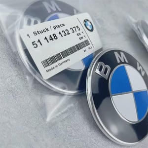 Könnte beinhalten: Schwarzes und silbernes BMW-Emblem mit dem BMW-Logo in Blau und Weiß. Ein Etikett lautet "1 Stuck / piece" und "51 148 132 375" mit dem Text "Made in Germany". Das Emblem ist rund und glänzend.
