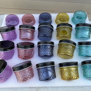 May include: A collection of small, decorative glass jars in various colors, including purple, red, blue, yellow, and teal. Each jar has a diamond pattern and a black lid. The jars are arranged on a white surface.