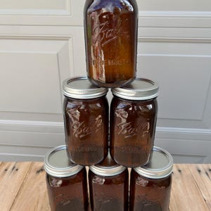 May include: A pyramid stack of six amber-colored glass jars with silver metal lids. Each jar features the embossed Ball logo and the text WIDE MOUTH on the front surface. The jars are arranged in a triangular formation with one on top.