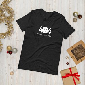 Puede incluir: Camiseta gris oscuro con el número 404 y un gráfico de cara triste en blanco. El texto "OH OH, WHAT NOW?" está debajo. La camiseta se muestra sobre una superficie de madera clara con adornos navideños.