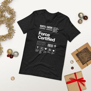 Puede incluir: Una camiseta gris oscuro con el texto "100% GEEK Organic Approved Force Certified" en blanco. La camiseta presenta un código de barras e iconos. La camiseta está doblada, con decoraciones navideñas.