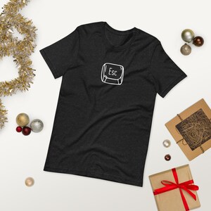 Puede incluir: Camiseta gris oscuro con el contorno blanco de la tecla "Esc". La camiseta está rodeada de adornos navideños, incluyendo adornos dorados y plateados y regalos envueltos.