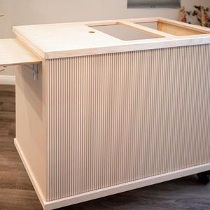May include: A light-coloured wooden kitchen island with a fluted design on the sides. The island has a countertop with a recessed area and a pull-out extension. It sits on wheels for mobility.