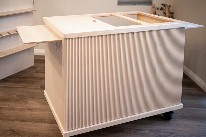 May include: A light-coloured wooden kitchen island with a fluted design on the sides. The island has a countertop with a recessed area and a pull-out extension. It sits on wheels for mobility.
