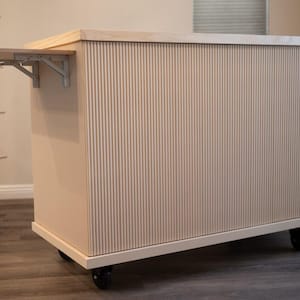 May include: A light-coloured, wooden kitchen island with a vertical grooved design. It features a fold-out shelf supported by metal brackets and rolls on black caster wheels. The island has a rectangular shape and a light wood countertop.