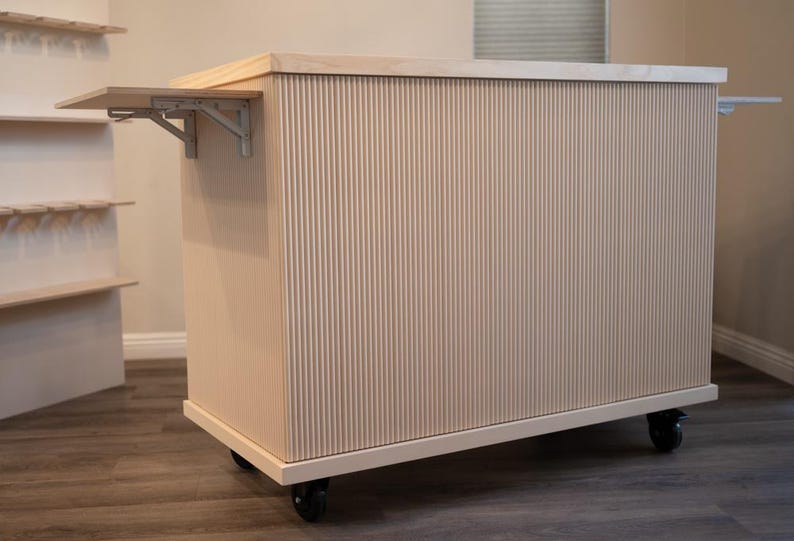 May include: A light-coloured, wooden kitchen island with a vertical grooved design. It features a fold-out shelf supported by metal brackets and rolls on black caster wheels. The island has a rectangular shape and a light wood countertop.