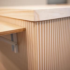 May include: Close-up of a light-coloured wooden cabinet with a fold-out shelf. The cabinet has vertical grooved detailing on the sides and a smooth, flat top. The shelf is supported by a white metal bracket.