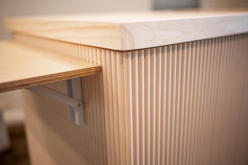 May include: Close-up of a light-coloured wooden cabinet with a fold-out shelf. The cabinet has vertical grooved detailing on the sides and a smooth, flat top. The shelf is supported by a white metal bracket.