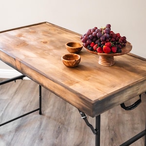 May include: A rectangular wooden table with a stained finish and a raised edge, supported by black metal folding legs. On the surface sit two small wooden bowls and a wooden pedestal bowl filled with red strawberries, blackberries, and grapes.