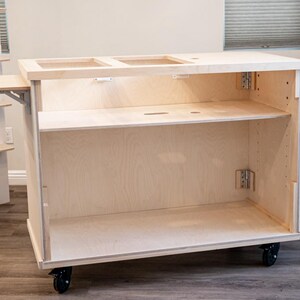 May include: A light-coloured wooden craft cart with multiple shelves and a fold-out side table. The cart has a rectangular shape and sits on black caster wheels. The interior features open shelving and cutouts on the top surface.