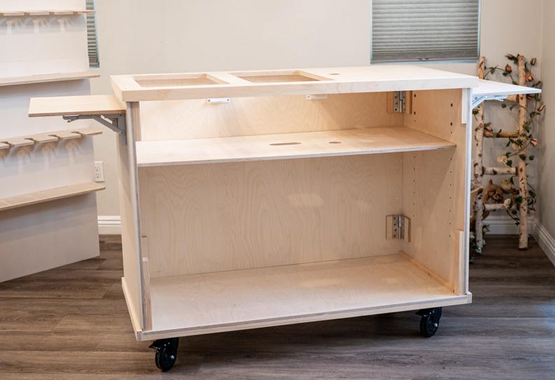 May include: A light-coloured wooden craft cart with multiple shelves and a fold-out side table. The cart has a rectangular shape and sits on black caster wheels. The interior features open shelving and cutouts on the top surface.