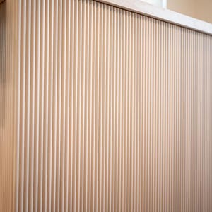 May include: A light-coloured, vertically-slatted wooden cabinet or counter. The design features closely spaced, parallel grooves, creating a textured surface. The top edge is a smooth, light-coloured surface. A metal bracket is visible on the left side.