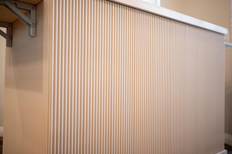 May include: A light-coloured, vertically-slatted wooden cabinet or counter. The design features closely spaced, parallel grooves, creating a textured surface. The top edge is a smooth, light-coloured surface. A metal bracket is visible on the left side.