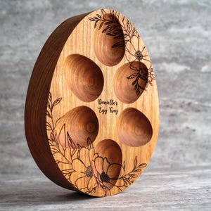 Peut inclure: Un plateau &agrave; &oelig;ufs en bois en forme d'&oelig;uf avec cinq indentations pour les &oelig;ufs. Le plateau est d&eacute;cor&eacute; d'un motif floral et du texte "Danielle's Egg Tray".