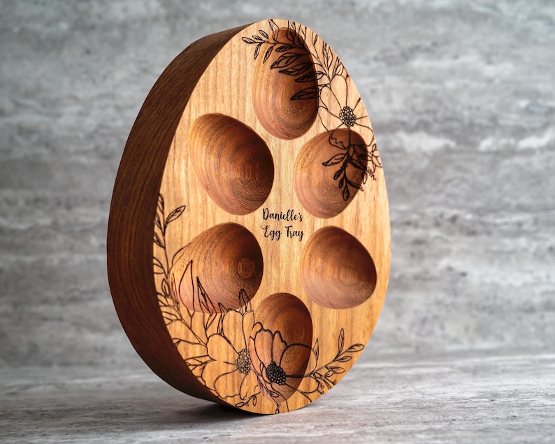 Peut inclure: Un plateau &agrave; &oelig;ufs en bois en forme d'&oelig;uf avec cinq indentations pour les &oelig;ufs. Le plateau est d&eacute;cor&eacute; d'un motif floral et du texte "Danielle's Egg Tray".