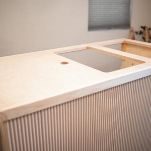 May include: A light-coloured wooden bar with a grooved front panel. The bar has cutouts for a sink and other appliances. A small, fold-out table is attached to the side. The bar is unfinished.