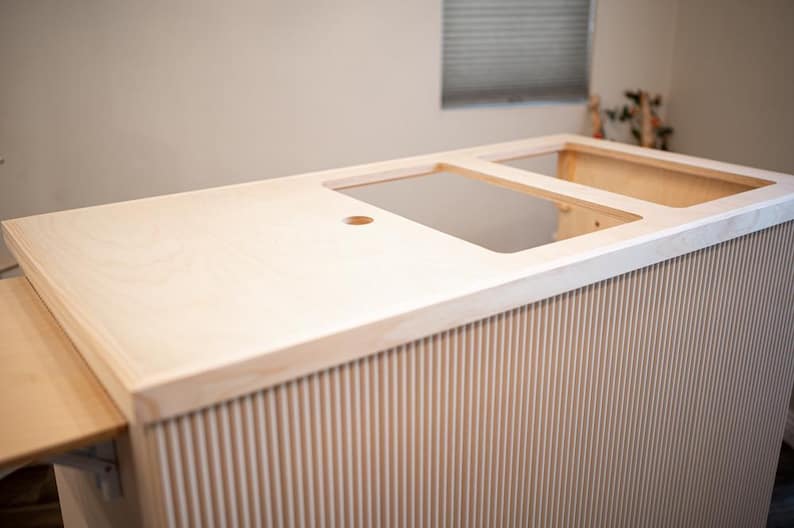May include: A light-coloured wooden bar with a grooved front panel. The bar has cutouts for a sink and other appliances. A small, fold-out table is attached to the side. The bar is unfinished.
