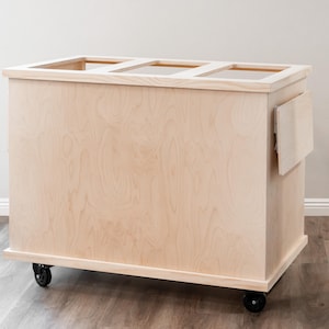 K&ouml;nnte beinhalten: Ein rollender Wagen aus Naturholz mit schlichtem Design. Der Wagen hat drei offene Regalb&ouml;den und ein kleines Seitenregal. Der Wagen ist aus unbehandeltem Holz und hat schwarze R&auml;der.