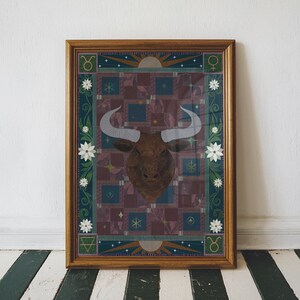 May include: A framed print of a brown bull's head with a geometric pattern background. The print features a stylised bull's head with white horns and a dark brown face. The background is a patchwork of squares in shades of purple, blue, and green, with gold stars and other decorative elements.