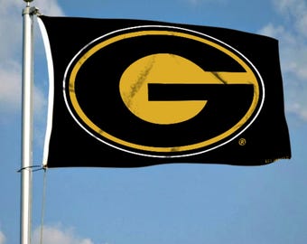 May include: A black flag with a white circle and a gold letter G in the center. The flag is flying on a flagpole against a blue sky.