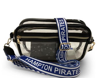 May include: A clear rectangular crossbody bag with black trim and gold-tone hardware. The bag features a blue and white adjustable strap with the text HAMPTON PIRATES printed repeatedly in white. A black zippered pocket is visible on the front.