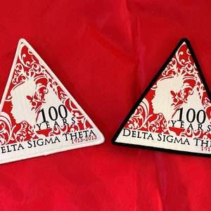 May include: Two triangular patches with black and white borders. The patches are red with white lettering and a white silhouette of two women's heads. The text on the patches reads "100 Years Delta Sigma Theta 1913-2013".