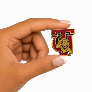 May include: A maroon and gold enamel pin shaped like the letter T, featuring a detailed graphic of a snarling tiger in yellow and black stripes. The design includes a small TM symbol in the bottom right corner, with a metallic gold border.