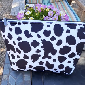 May include: A zippered pouch featuring a black and white cow print pattern. The rectangular bag has a black zipper closure with a metallic pull tab. The fabric displays an irregular spotted design across the entire front surface of the item.