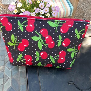 May include: A black zippered pouch featuring a repeating pattern of red cherries with green leaves and stems against a background of small white polka dots. The pouch has a red zipper closure along the top edge and a rectangular, flat-bottomed shape.