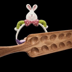 May include: A wooden egg holder with a handle and 15 indentations for eggs. The holder is brown and has a natural wood finish. A ceramic Easter bunny with a pink bow tie is in the background.