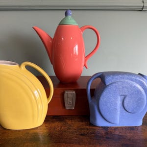 May include: A collection of colorful ceramic teapots and pitchers. The set includes a yellow pitcher, a coral-colored teapot with a green lid, and a blue pitcher. The items are displayed on a wooden surface.