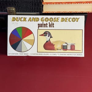 Puede incluir: Una caja vintage de "Duck and Goose Decoy paint kit". La caja presenta una rueda de color, una ilustración de pato pintado y el texto "paint kit". El kit incluye un pincel, 12 colores mezclados y un manual de instrucciones de pintura.