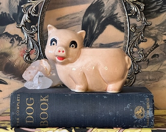 Puede incluir: Una figurita de cerdo de cerámica rosa con ojos negros y una nariz rosa. El cerdo está sentado encima de dos libros, uno con el título "The Complete Dog Book" y el otro con el título "The Flowering of Art Nouveau".