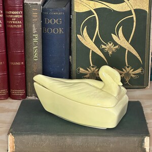 Può includere: Un piatto in ceramica giallo a forma di cigno con un coperchio. Il piatto è appoggiato su un libro con il titolo "Art Appreciation" sul dorso.