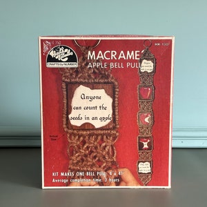 May include: A box for a macrame apple bell pull craft kit. The box is red with a picture of the finished project, which is a brown macrame bell pull with a wooden frame that says "Anyone can count the seeds in an apple". The box also includes the text "HK 130" and "Crafts by Number".