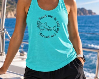 May include: A bright turquoise racerback tank top featuring a black graphic of a shark in the center, surrounded by the circular text "Feed me & tell me I'm pretty." The tank top is paired with black athletic shorts, creating a casual and sporty look.