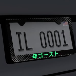 May include: A black carbon fiber license plate frame with a white license plate that reads "IL 0001". The frame has a small American flag sticker in the top right corner and a green glow-in-the-dark sticker with Japanese text that reads "ゴースト" on the bottom.