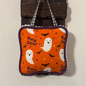 May include: Halloween-themed decorative pillow with an orange background featuring white ghosts, bats, and stars. The pillow has a purple border and white pom-pom trim. The handle is black and white checkered.