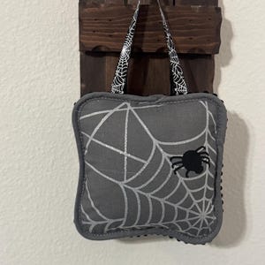 May include: A gray Halloween-themed pillow with a spiderweb design and a black spider. The pillow is hanging from a dark wooden plaque with a black ribbon featuring a spiderweb pattern. The pillow is a decorative item.