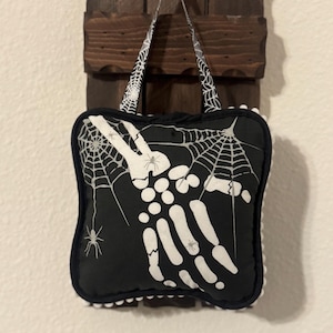 May include: A black and white Halloween-themed tote bag. The bag features a skeleton hand design with spiderwebs and spiders. The bag has a white pom-pom trim and a handle with a spiderweb pattern. The bag is hanging on a wooden wall decor.