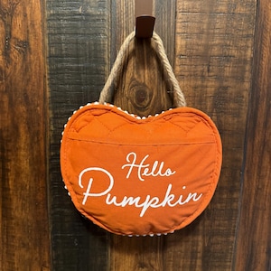 May include: Orange heart-shaped door hanger with white trim and the text "Hello Pumpkin" in white.