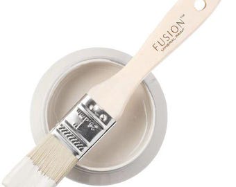 May include: A paint brush with a light-colored wooden handle featuring the text FUSION MINERAL PAINT and a metal ferrule stamped with 1 inch and 25.4mm, resting across a round container filled with light gray paint and white bristles.