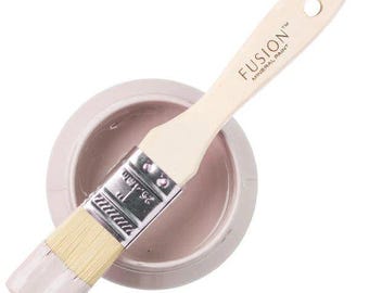 May include: A light pink paint brush with a wooden handle labeled Fusion Mineral Paint resting across a round container filled with matching light pink paint. The metal ferrule of the brush is clearly marked with the measurement 1 inch or 25.4 mm.