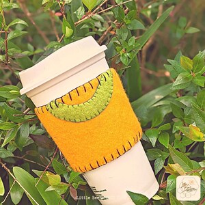 Könnte beinhalten: Ein gelber Filz-Becherwärmer mit einem grünen Filz-Halbmond-Design und schwarzen Nähten. Der Wärmer befindet sich auf einem weißen To-Go-Becher.