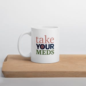 May include: White ceramic mug with the words "take YOUR MEDS" in a stacked design. The word "take" is in a dusty rose color, "YOUR" is navy blue with a brain graphic, and "MEDS" is olive green. The mug sits on a wooden surface.