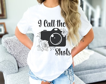 May include: White t-shirt with black text that reads "I Call the Shots" and a black silhouette of a camera with white floral accents.