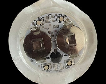 May include: Close-up of a round, white circuit board with two silver battery compartments labeled "BAT1" and "BAT2". The board has six LEDs labeled "LED4", "LED5", "LED6", "LED3", "LED2", and "LED1".